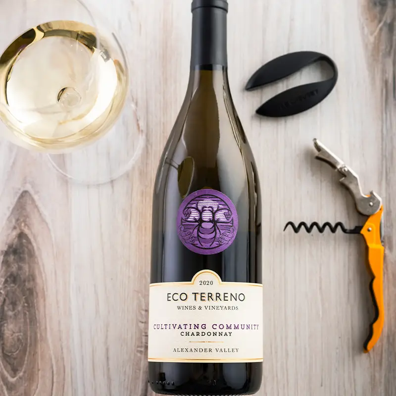 Bottle of Eco Terreno Cultivating Community Chardonnay lying on a wooden table with a glass of white wine and a corkscrew next to it.