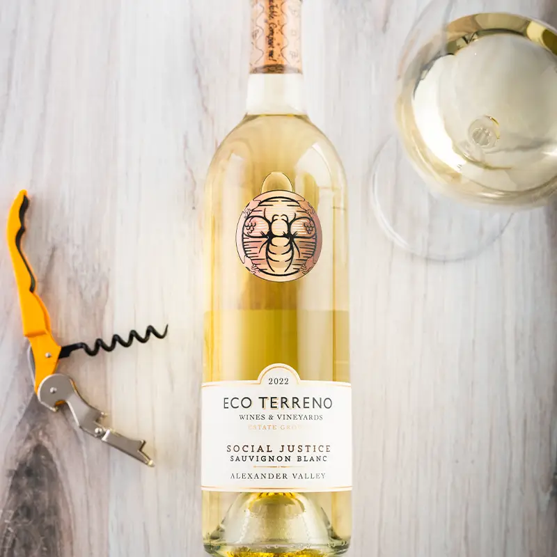 Bottle of Eco Terreno Social Justice Sauvignon Blanc lying on a wooden table with a glass of white wine and a corkscrew next to it.